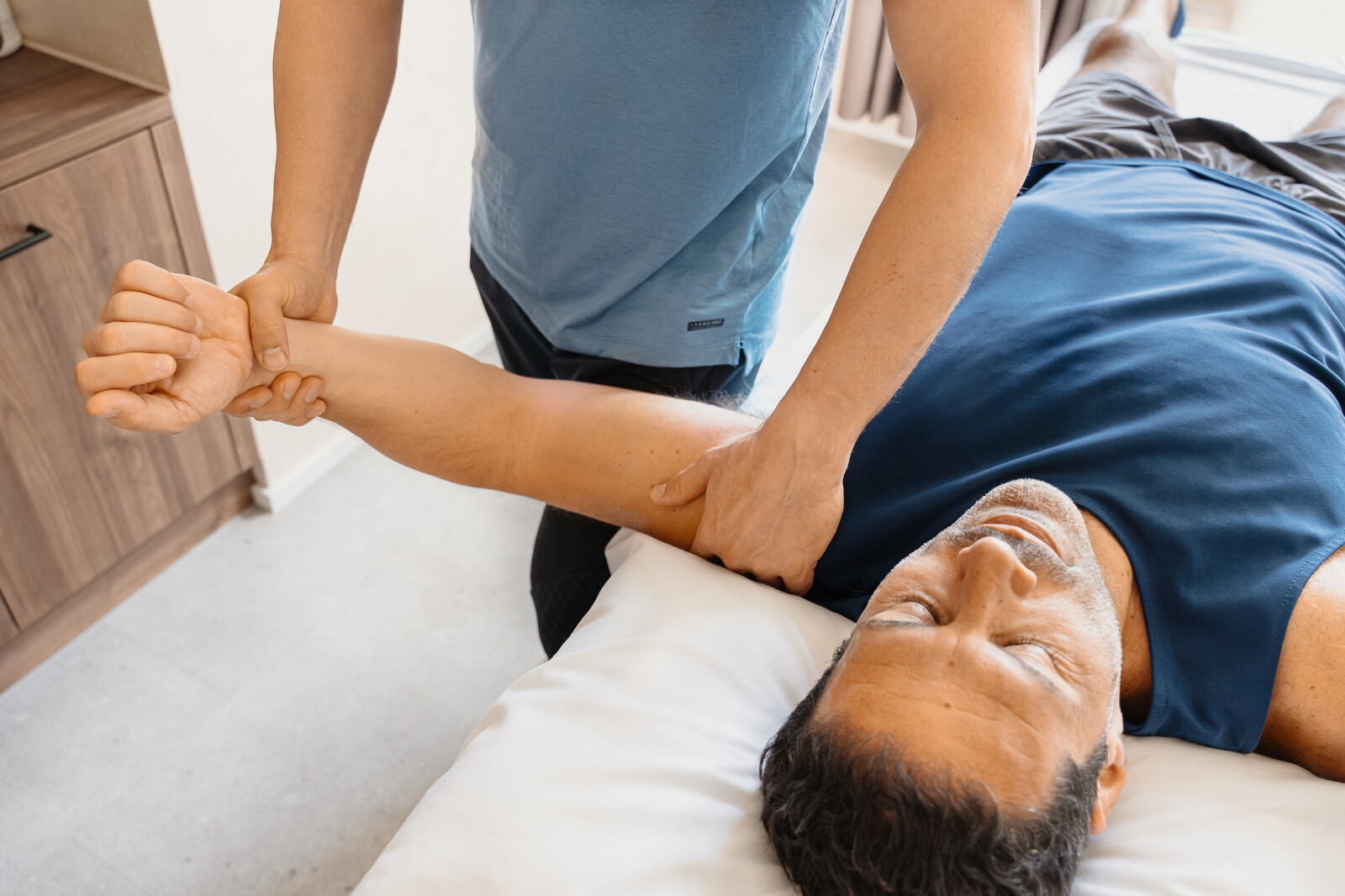 Eine Person erhält eine Armtherapie, während sie auf einem Massagetisch liegt. Ein Therapeut in einem blauen Hemd manipuliert sanft den entspannten Arm des Patienten. Ruhige Umgebung.