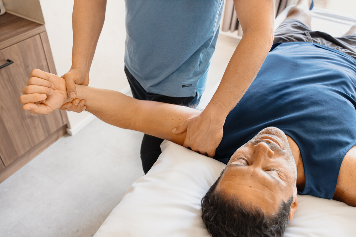 Eine Person erhält eine Armtherapie, während sie auf einem Massagetisch liegt. Ein Therapeut in einem blauen Hemd manipuliert sanft den entspannten Arm des Patienten. Ruhige Umgebung.