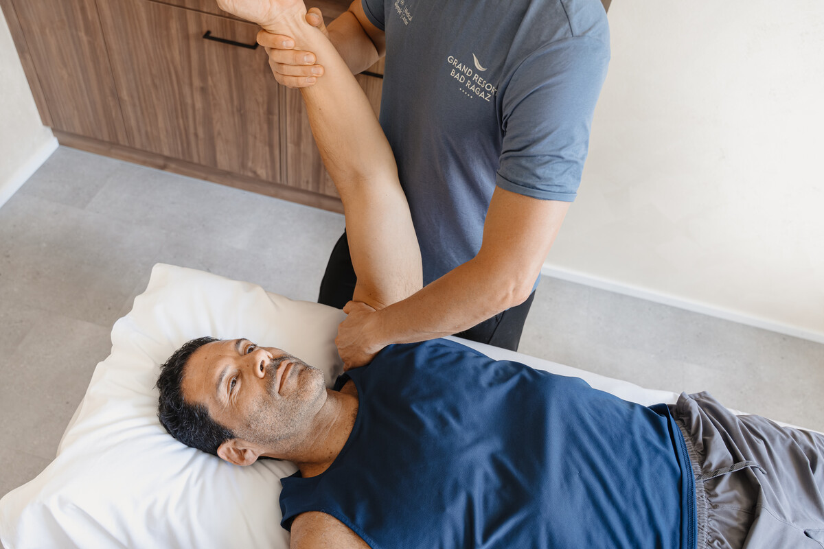 Ein Mann liegt auf einem Bett und erhält Physiotherapie. Der Therapeut, der ein blaues Hemd trägt, dehnt sanft den Arm des Patienten. Die Umgebung ist ruhig und klinisch.
