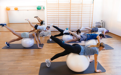 Ein Gruppenkurs mit sieben Teilnehmern, die auf Fitnessbällen balancieren und auf Matten in einem hellen Fitnessstudio Beinheben machen. Die Atmosphäre ist konzentriert und energiegeladen.