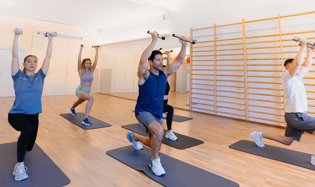 Ein Gruppenkurs mit vier Teilnehmern in einem hellen Studio, die mit über dem Kopf erhobenen Hanteln Ausfallschritte machen. Die Stimmung ist energiegeladen und konzentriert.