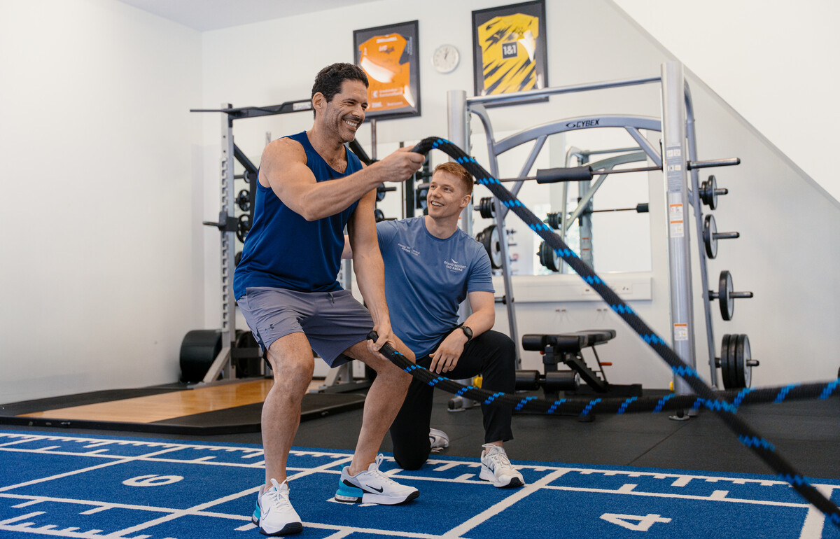 A man exercises with battle ropes in a gym, smiling, while a trainer kneels beside him, offering encouragement. The setting is energetic and supportive.