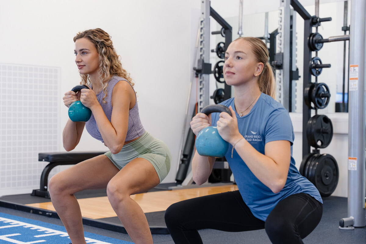 Zwei Frauen machen in einem Fitnessstudio konzentriert und entschlossen Kettlebell-Kniebeugen. Sie halten blaue Kettlebells in den Händen, im Hintergrund sind Fitnessgeräte zu sehen.