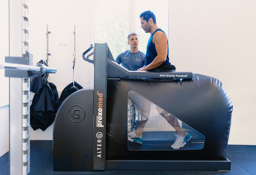 Ein Mann in Trainingskleidung benutzt unter Aufsicht eines Trainers in einem Fitnessstudio ein Anti-Schwerkraft-Laufband. Die Szene ist gut ausgeleuchtet und vermittelt Konzentration und Intensität.