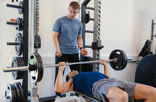 Ein Trainer leitet eine Person an, die in einem Fitnessstudio Bankdrücken macht. Der Trainer beobachtet aufmerksam, zeigt Konzentration und Unterstützung. Gewichte und Fitnessgeräte sind zu sehen.