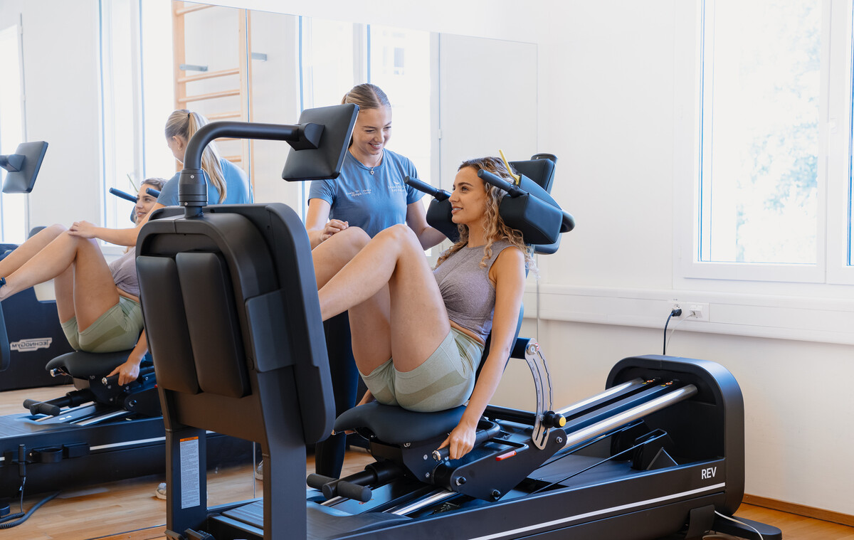 Eine Frau an einer Beinpresse lächelt einen Trainer in einem Fitnessstudio an. Sie befinden sich in einem hellen Raum mit Spiegeln und großen Fenstern, der eine positive, unterstützende Atmosphäre schafft.