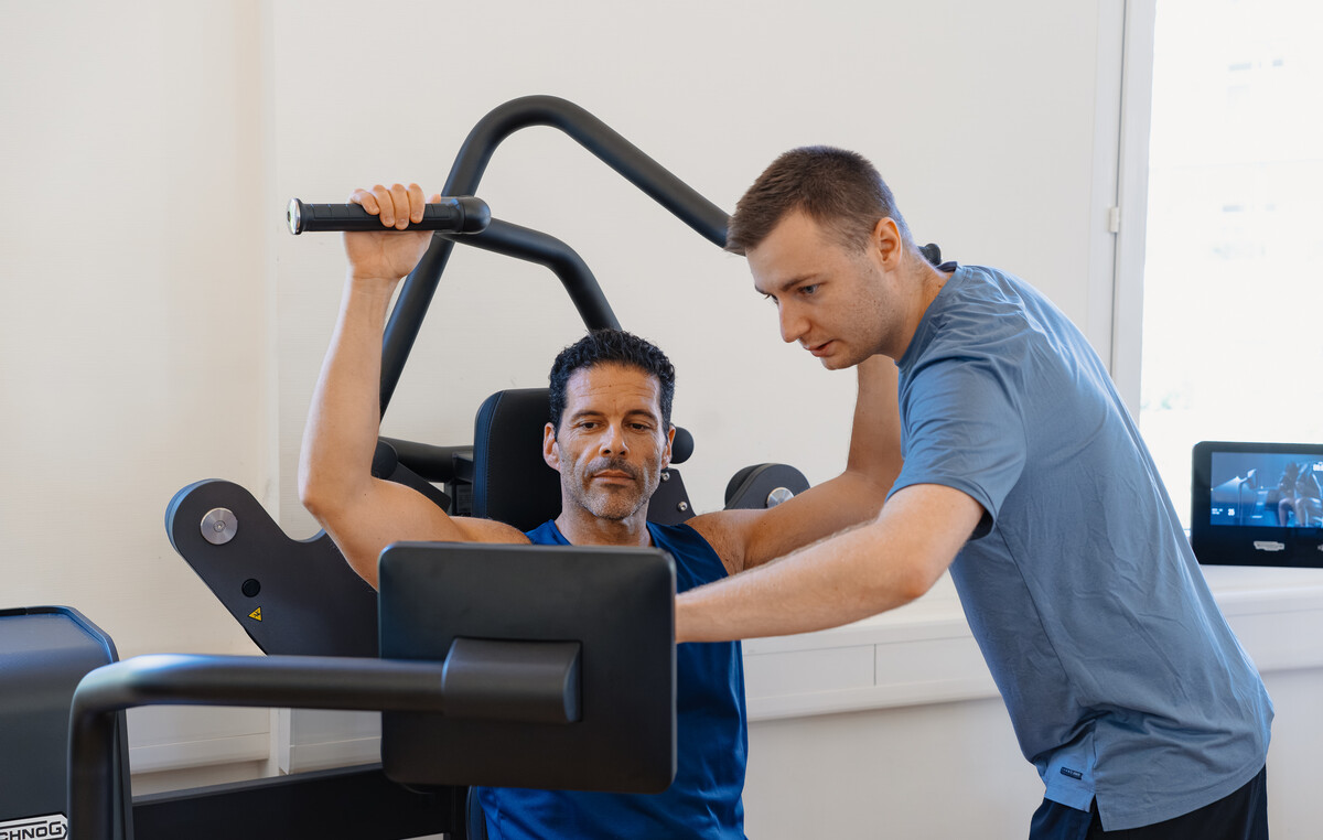 Ein Personal Trainer begleitet einen Mann an einem Kraftgerät in einem hellen Fitnessstudio. Der Mann ist konzentriert, während der Trainer ihm die richtige Ausführung zeigt. Beide tragen Sportbekleidung.