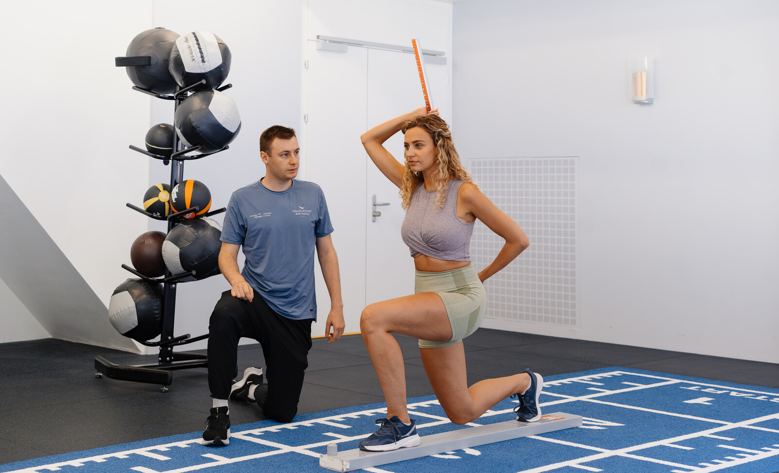Eine Frau in Sportbekleidung führt unter Anleitung eines Trainers eine Ausfallschritt-Bewertung mit einem Balance-Stab durch. Sie befinden sich in einem Fitnessstudio mit Gymnastikbällen im Hintergrund.