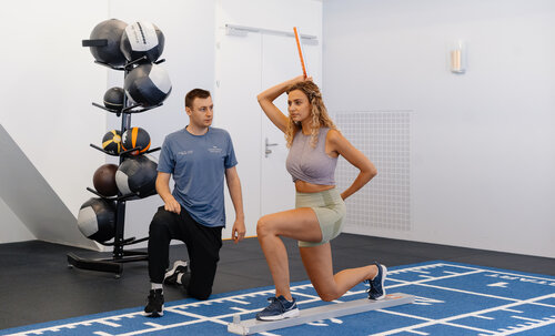 Eine Frau in Sportbekleidung führt unter Anleitung eines Trainers eine Ausfallschritt-Bewertung mit einem Balance-Stab durch. Sie befinden sich in einem Fitnessstudio mit Gymnastikbällen im Hintergrund.
