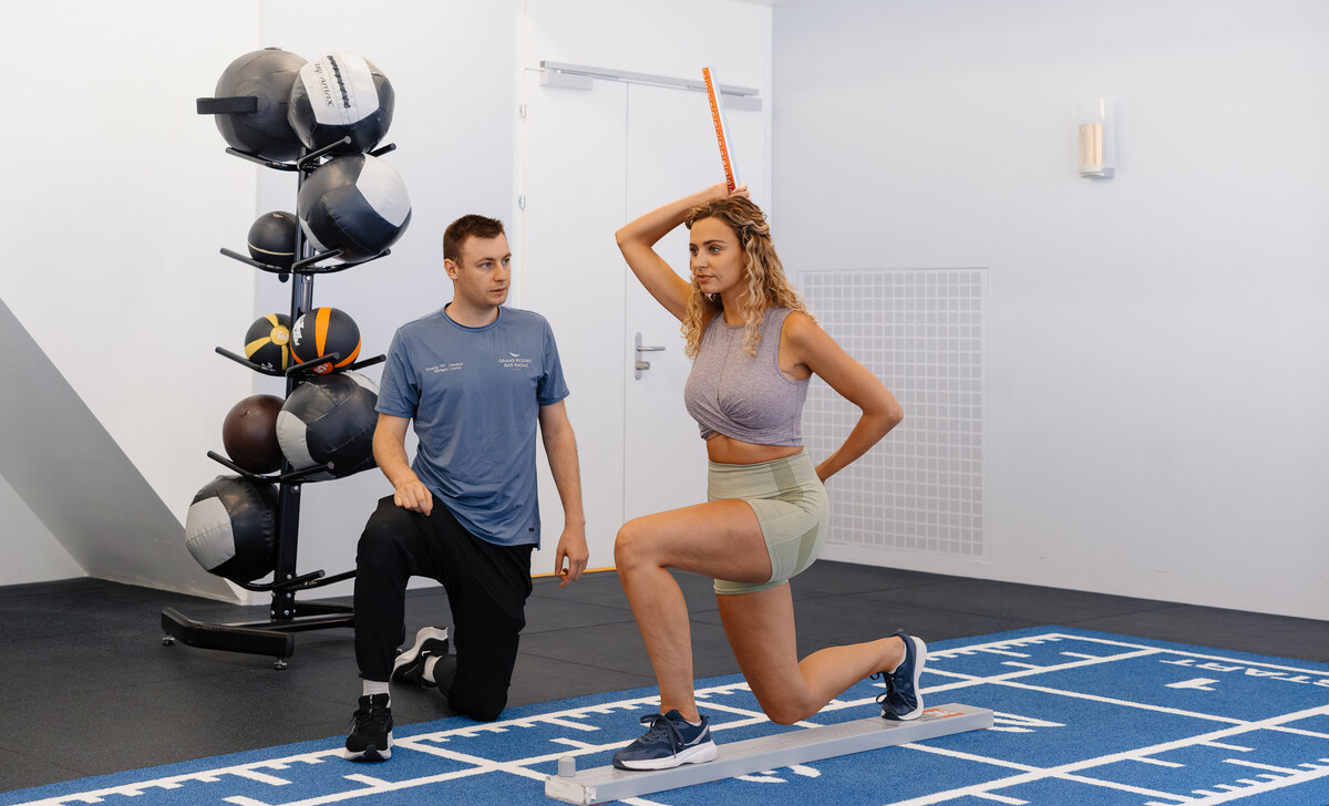 Eine Frau in Sportbekleidung führt unter Anleitung eines Trainers eine Ausfallschritt-Bewertung mit einem Balance-Stab durch. Sie befinden sich in einem Fitnessstudio mit Gymnastikbällen im Hintergrund.