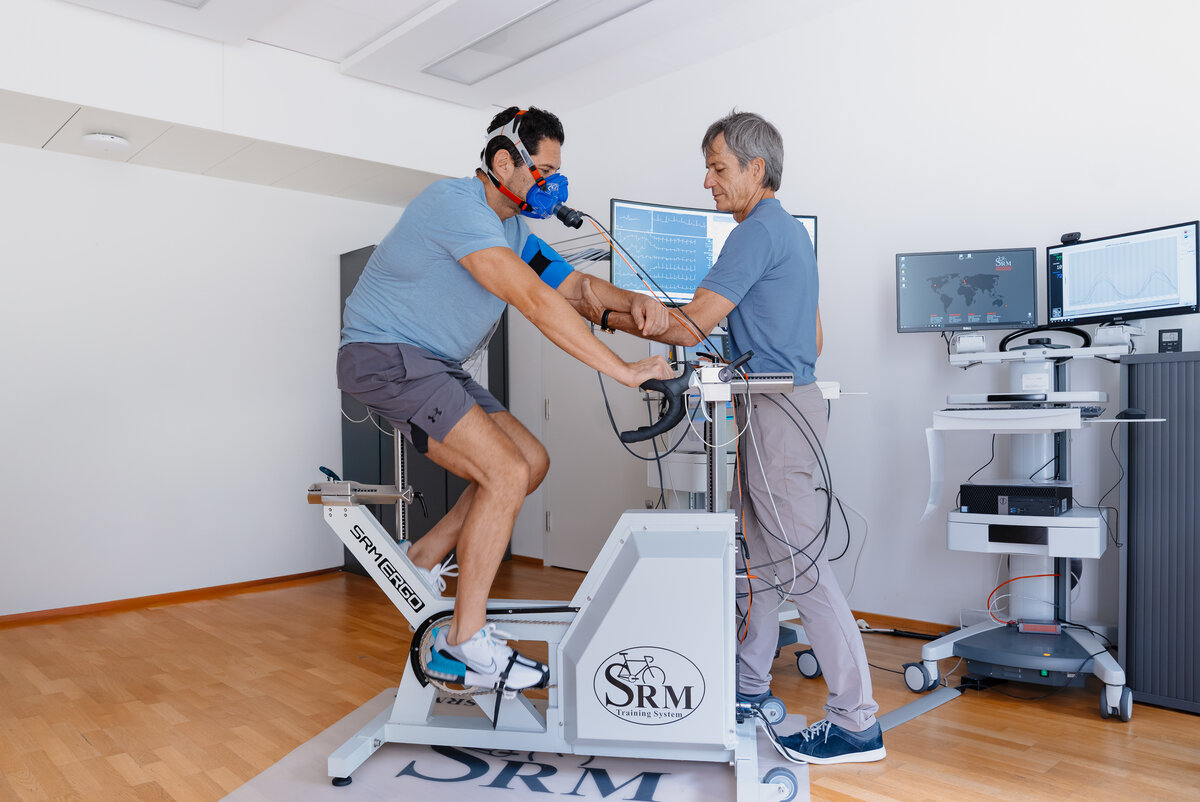 Ein Mann in Sportkleidung sitzt auf einem Hightech-Heimtrainer, trägt eine blaue Maske und Sensoren. Ein Trainer überwacht die Daten auf Bildschirmen in der Nähe in einer klinischen Umgebung.