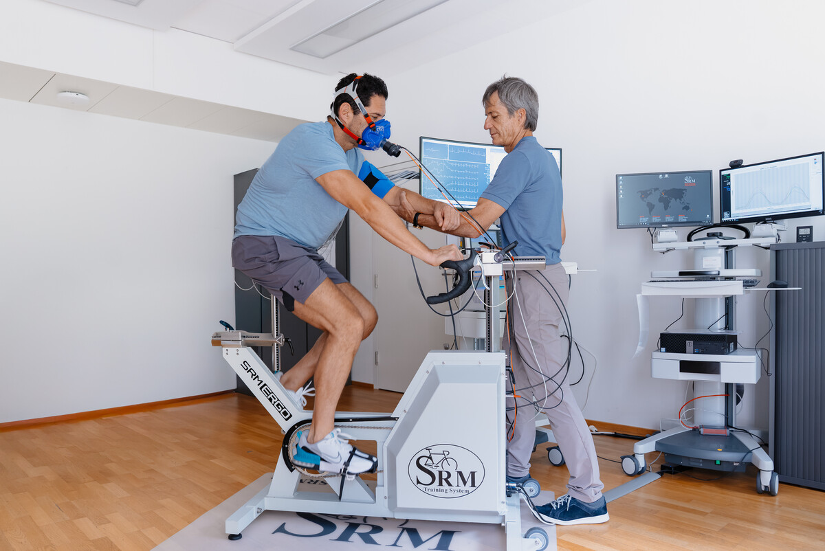 Ein Mann in Sportkleidung sitzt auf einem Hightech-Heimtrainer, trägt eine blaue Maske und Sensoren. Ein Trainer überwacht die Daten auf Bildschirmen in der Nähe in einer klinischen Umgebung.