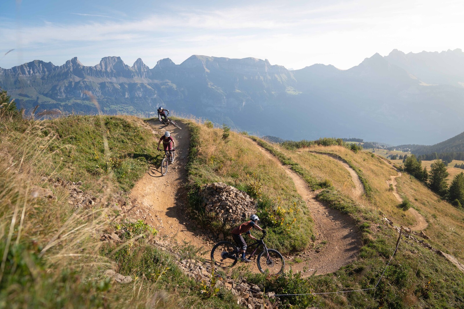 Mountainbiker fahren auf einem gewundenen Feldweg an einem grasbewachsenen Hang mit einer malerischen Bergkette im Hintergrund unter einem strahlend blauen Himmel und vermitteln ein Gefühl von Abenteuer.