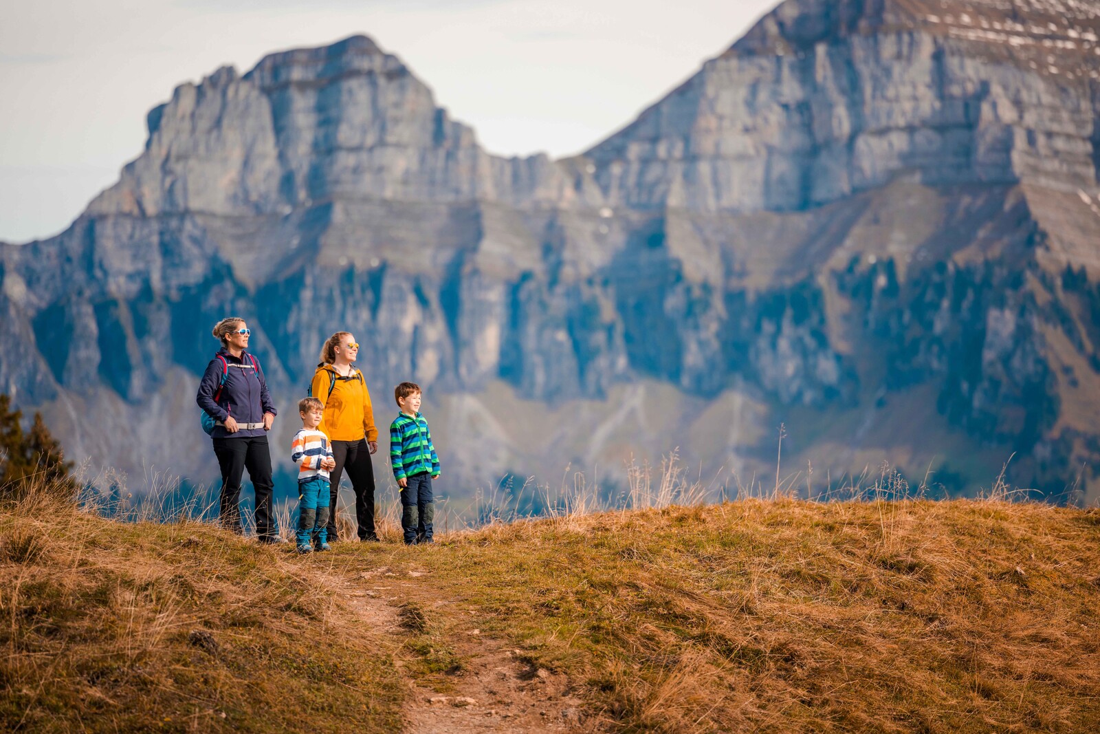 Eine vierköpfige Familie steht auf einem grasbewachsenen Hügel, alle tragen Jacken, zwei Kinder haben gestreifte Hemden an. Im Hintergrund erheben sich majestätische Felsberge unter einem klaren Himmel.