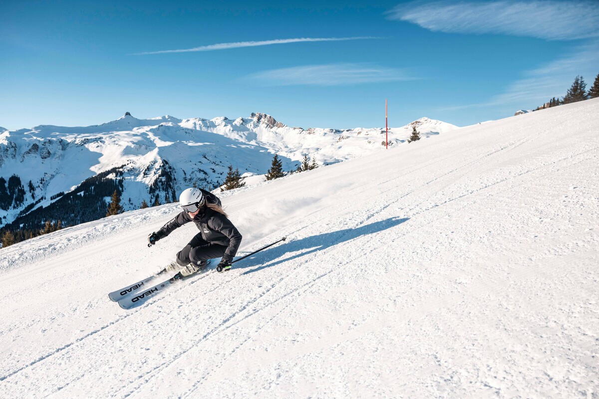 Ein Skifahrer in schwarzer Ausrüstung carvt einen sonnenbeschienenen, verschneiten Hang hinunter. Majestätische schneebedeckte Berge und ein strahlend blauer Himmel bilden die Kulisse und vermitteln ein Gefühl der Begeisterung.