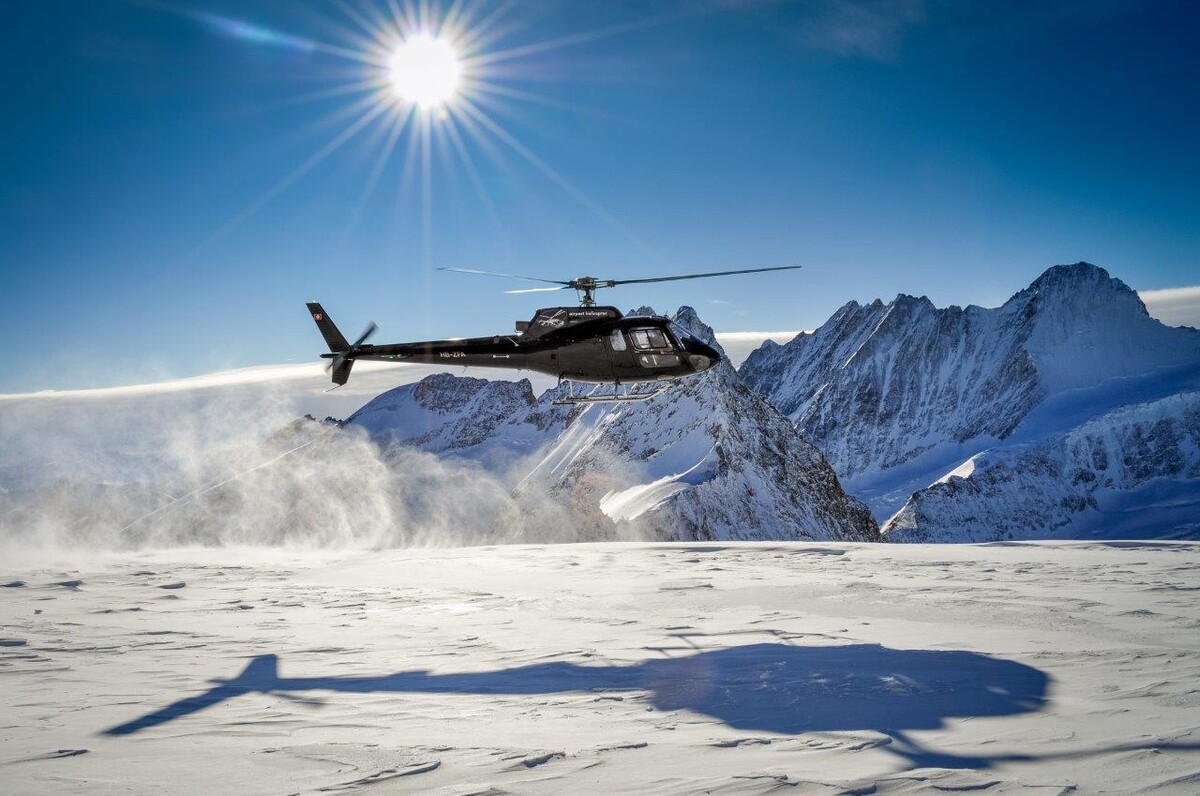 Ein schwarzer Helikopter der über schneebedeckte Berge fliegt während die Sonne strahlt.