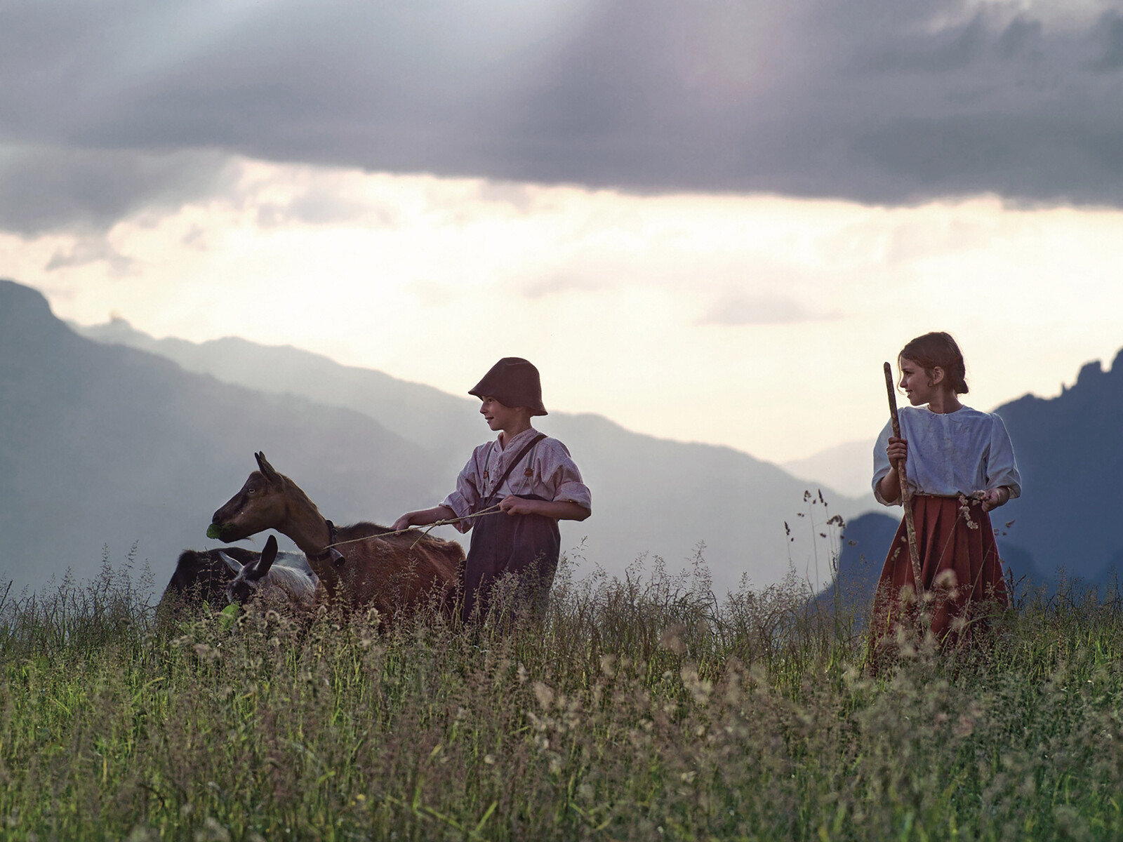 Heidi und Peter laufen mit einer Ziege über eine blühende Wiese in den Bergen.