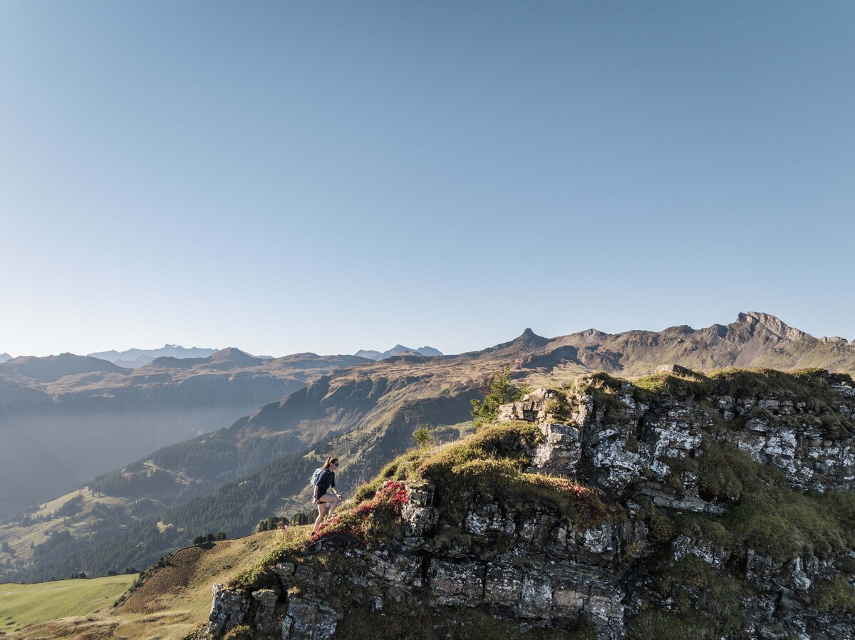 Ein Wanderer steht auf einem Felsvorsprung und blickt unter einem strahlend blauen Himmel auf eine weitläufige Bergkette der 7-Gipfel-Tour. Die Szene vermittelt ein Gefühl von Abenteuer und Ruhe.