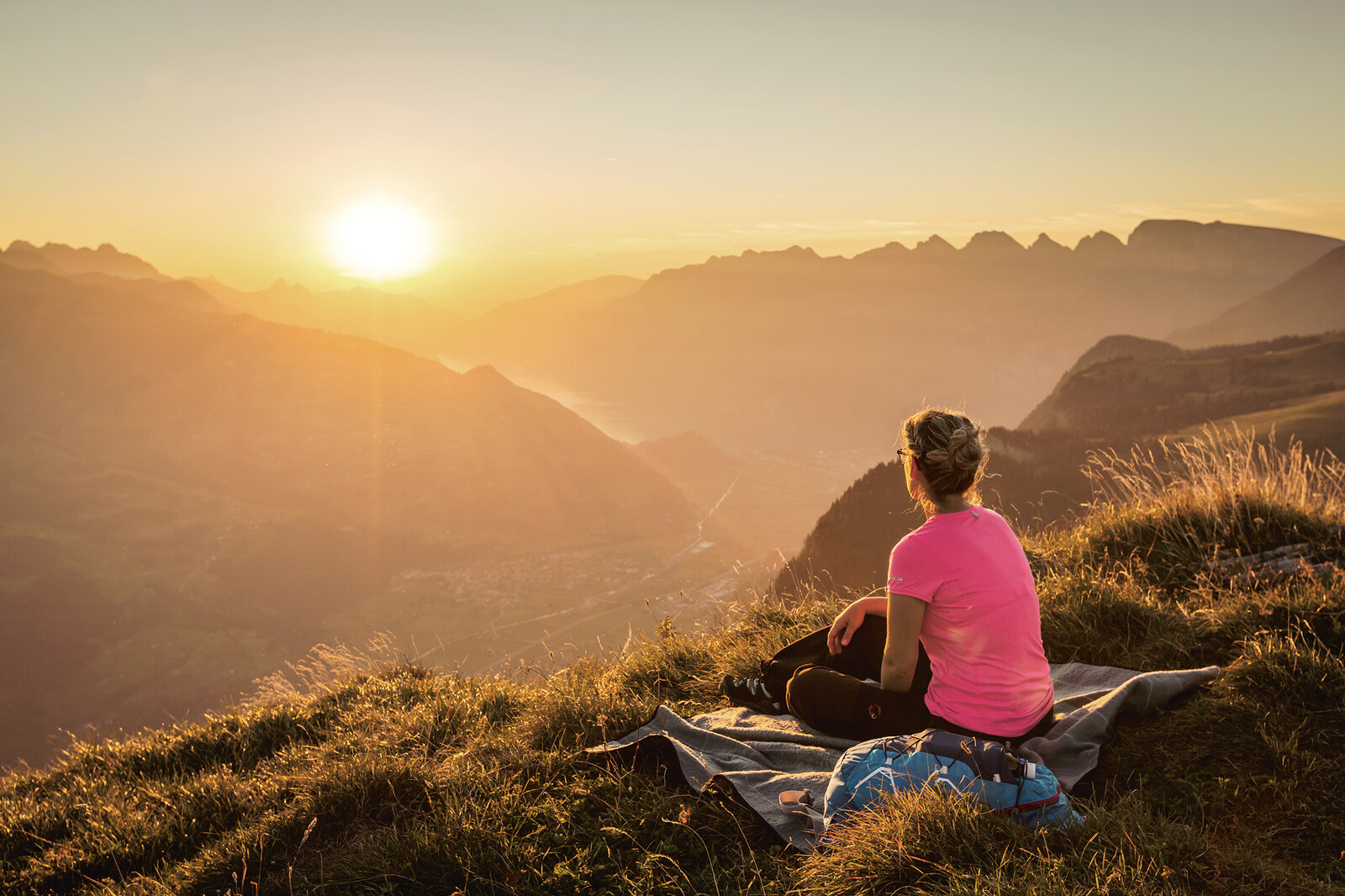 A woman sits on a blanket in the meadow and admires the sunset behind the mountains. The grass and the other mountains around them are bathed in the orange-gold of the setting sun.
