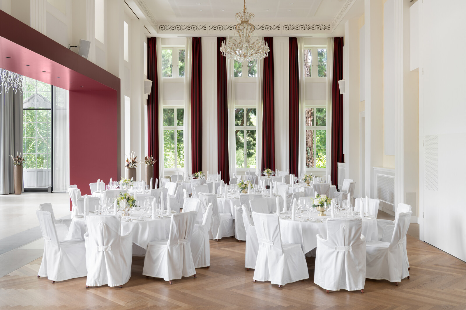 Der elegante Bernahrd Simon Saal mit grossen Fenstern und roten Vorhängen, an denen runde Tische mit weißen Tüchern und Stühlen stehen. Ein großer Kronleuchter hängt über dem Saal und schafft ein formelles, luxuriöses Ambiente.