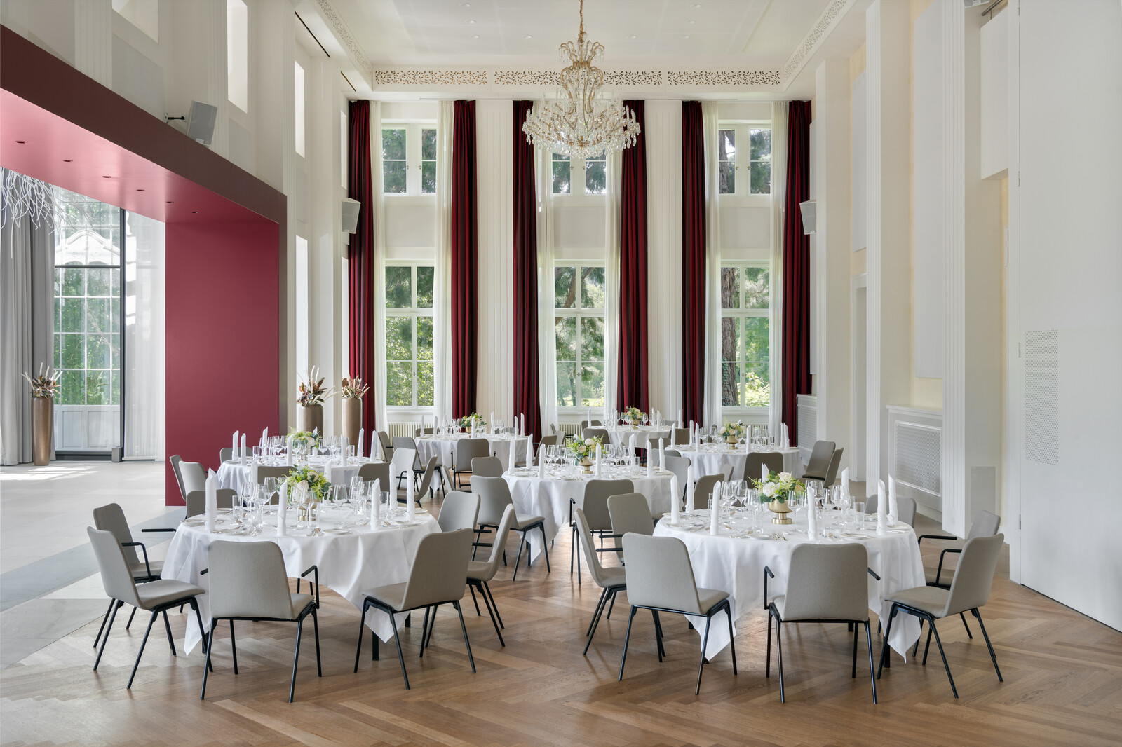 Eleganter Bernhard Simon Saal mit hohen Fenstern, roten Vorhängen und einem Kronleuchter. Runde Tische, gedeckt mit weißer Bettwäsche, Kerzen und Blumenschmuck.