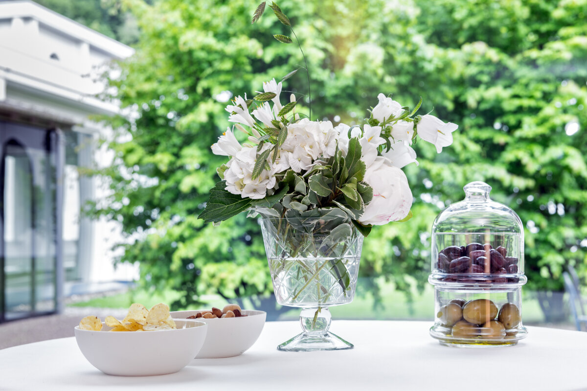 Elegant gedeckter Tisch mit einer Kristallvase mit weißen Blumen, Schalen mit Chips und Nüssen und einem Glas mit Oliven und Datteln, umgeben von üppigem Grün.
