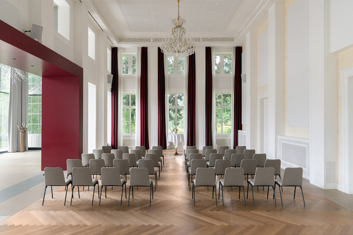 Elegant Bernhard Simon Hall with rows of gray chairs in front of high windows draped with burgundy curtains. A chandelier hangs from the ceiling. Bright and cheerful atmosphere.