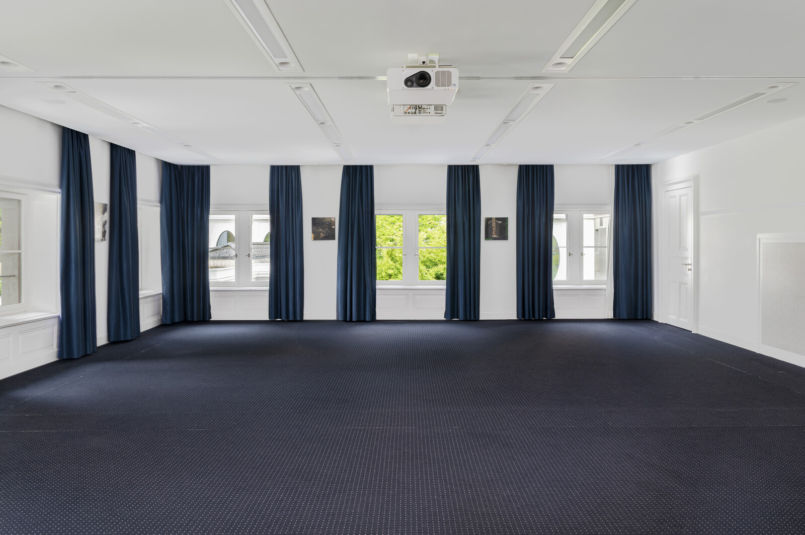 Der Johanna Spyri Saal mit dunkelblauem Teppich und passenden bodenlangen Vorhängen. Weiße Wände und große Fenster lassen natürliches Licht herein und vermitteln ein ruhiges Gefühl.
