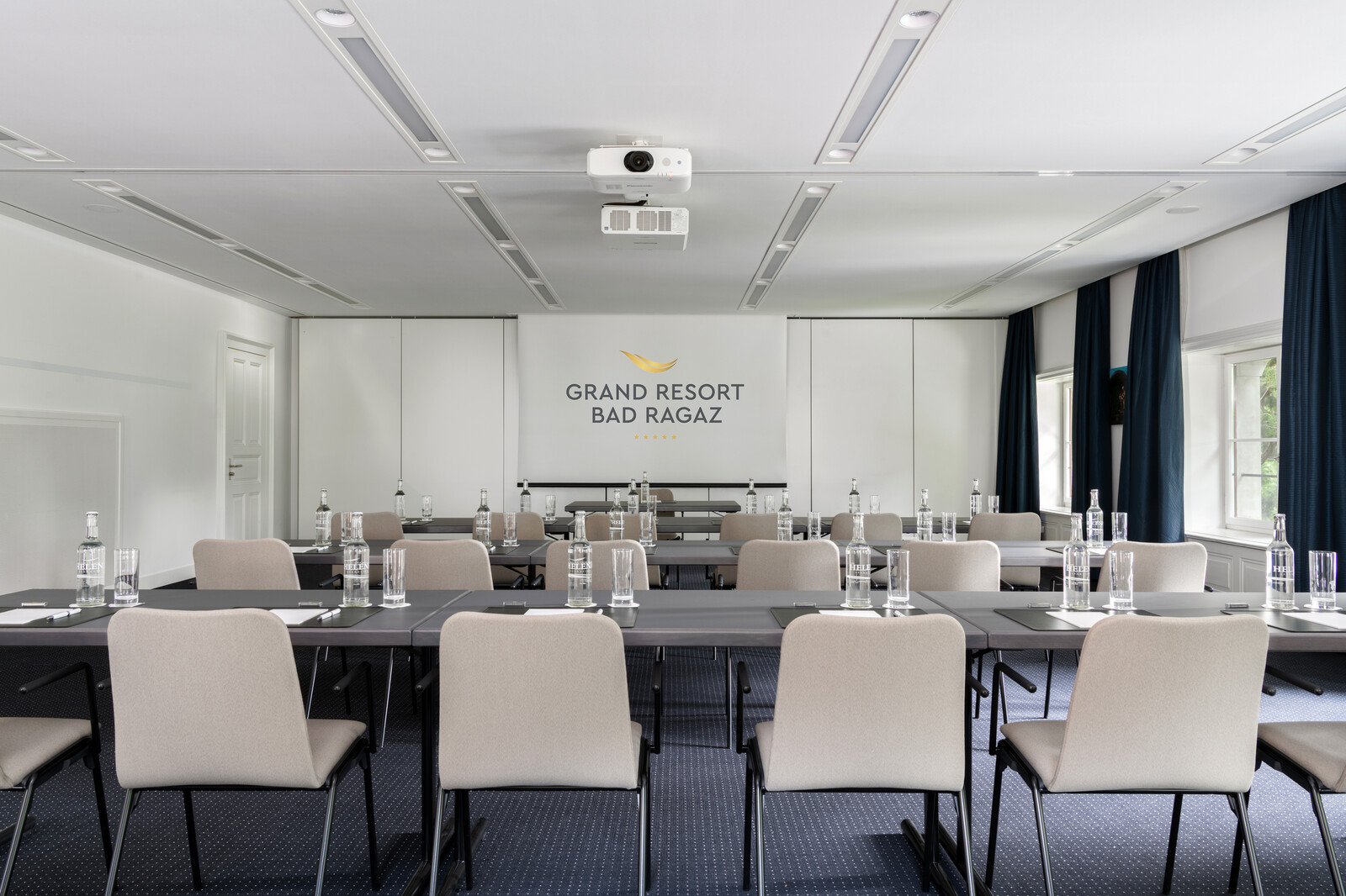 Der Johanna Spyri Saal im Kursaal des Hotel Grand Resort Bad Ragaz, einem Superior Luxushotel in der Schweiz, ist bereits für ein Meeting oder einen Workshop hergerichtet. Tische und Stühle stehen überall im Raum verteilt. An den Fenstern hängen dunkle Vorhänge, passend zu dem Teppich am Boden.