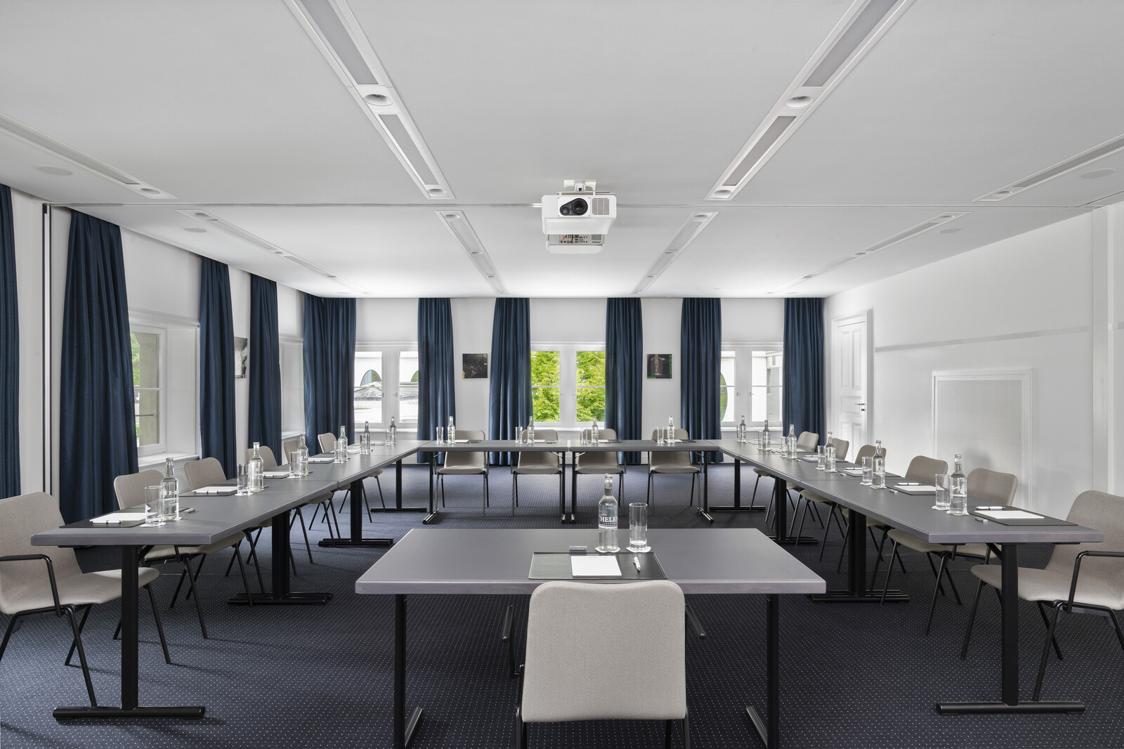 Der Johanna Spyri Saal im Kursaal des Hotel Grand Resort Bad Ragaz, einem Superior Luxushotel in der Schweiz, ist bereits für ein Meeting oder einen Workshop hergerichtet. Tische und Stühle stehen überall im Raum verteilt. An den Fenstern hängen dunkle Vorhänge, passend zu dem Teppich am Boden.