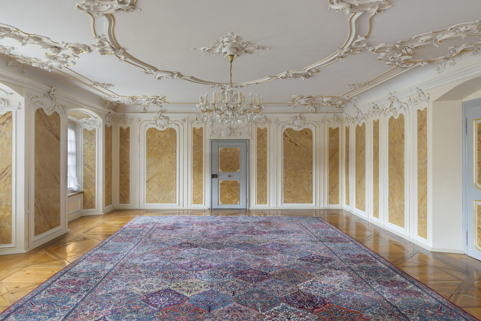 Der elegante Musiksalon mit kunstvoller Stuckdecke, Kronleuchter und gemustertem Perserteppich. Marmorwände und Parkettboden, die einen historischen, luxuriösen Ton vermitteln.