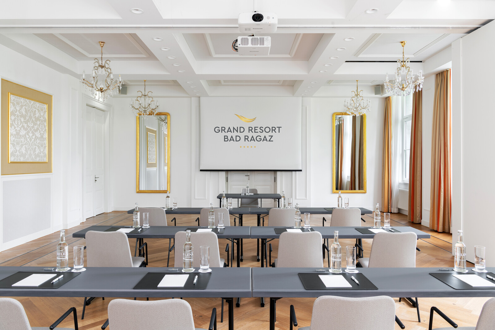 Der Rainer Maria Silke Saal im Grand Resort Bad Ragaz mit ordentlich aufgestellten Stühlen und Tischen, Notizblöcken, Stiften und Wasserflaschen. Elegante Kronleuchter und Spiegel sorgen für Raffinesse.