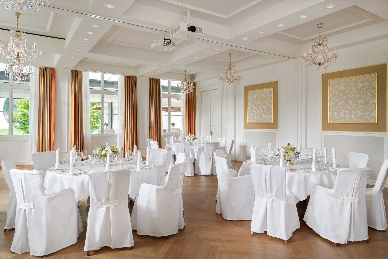 Der Rainer Maria Rilke Saal mit runden, weiss gedeckten Tischen, die mit Blumenkränzen geschmückt sind. Große Fenster, Kronleuchter und warme Beleuchtung sorgen für eine elegante Atmosphäre.