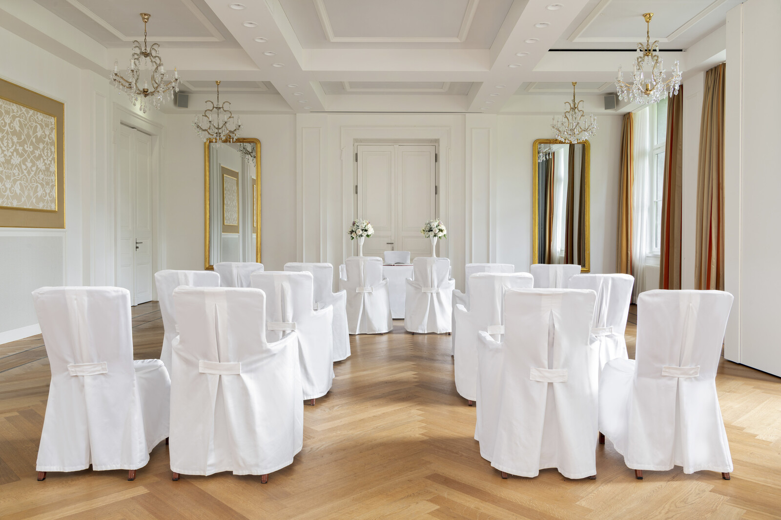 Eleganter Rainer Maria Rilke Saal mit weissen Stuhlhussen in halbkreisförmiger Anordnung. Kristallkronleuchter, goldgerahmte Spiegel und florale Akzente sorgen für ein heiteres Ambiente.