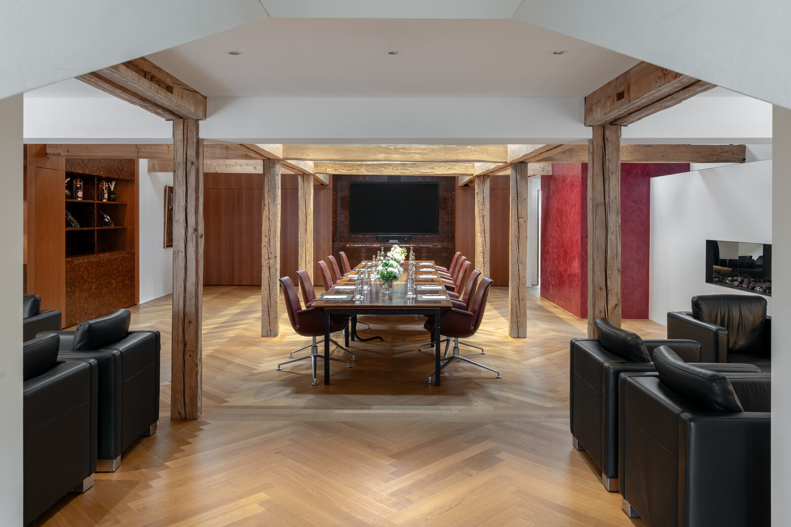 Die Hugo Lounge im Kursaal des Hotel Grand Resort Bad Ragaz, einem Superior Luxushotel in der Schweiz, ist bereits aufgedeckt und bereit für eine Sitzung. Die Wand auf der linken Seite ist weiss gestrichen, während die rechte Wand rot ist. In der Mitte des Raums steht ein hölzerner Tisch mit Stühlen. Ein Fernseher hängt an der Wand am Ende des Tisches. Holzbalken stützen das Dach.