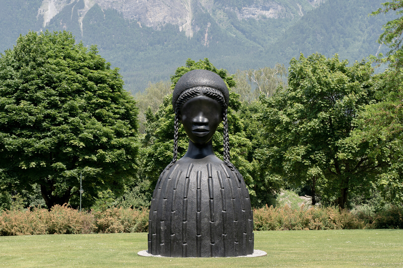Große schwarze Skulptur eines Frauenkopfes mit Zöpfen in einem Park. Üppig grüne Bäume und Berge im Hintergrund, die eine heitere Atmosphäre schaffen.