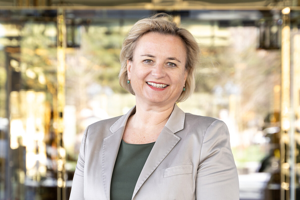 Eine lächelnde Person in Geschäftskleidung steht selbstbewusst im Freien, umrahmt von weichzeichnenden Grünflächen und Glasgebäuden, die eine professionelle Atmosphäre vermitteln.