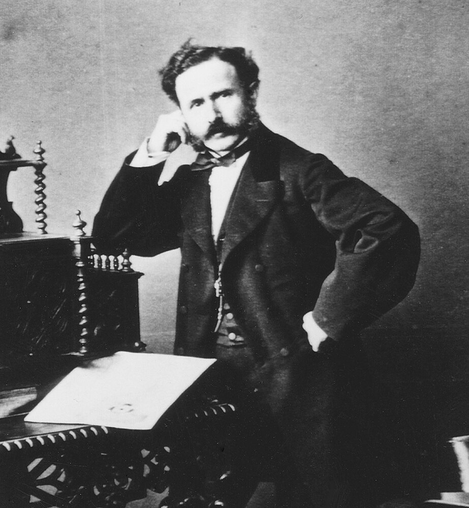 Ein Mann aus dem 19. Jahrhundert steht in nachdenklicher Pose neben einem verzierten Schreibtisch und stützt eine Hand auf sein Gesicht, was einen nachdenklichen und vornehmen Ton vermittelt.