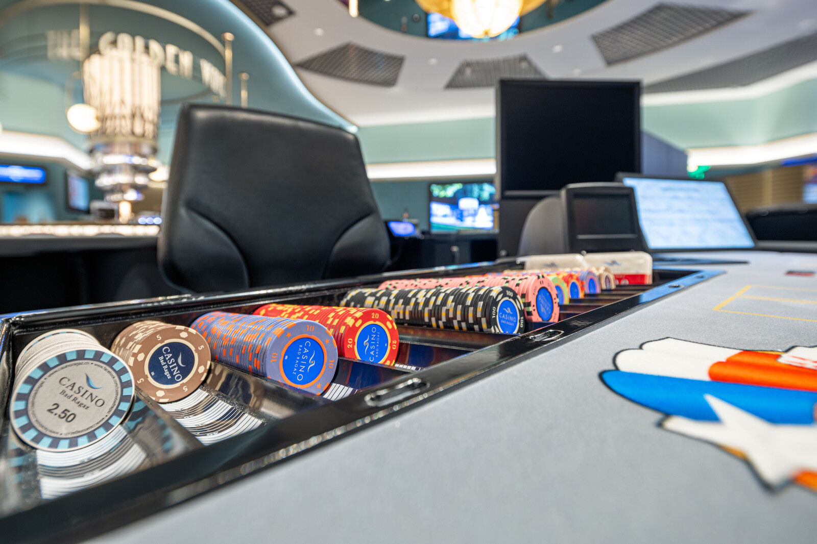 Pokerchips in verschiedenen Farben, die ordentlich auf einem Casinotisch gestapelt sind. In der modernen, gut beleuchteten Umgebung sind der Stuhl des Croupiers und elektronische Bildschirme zu sehen.