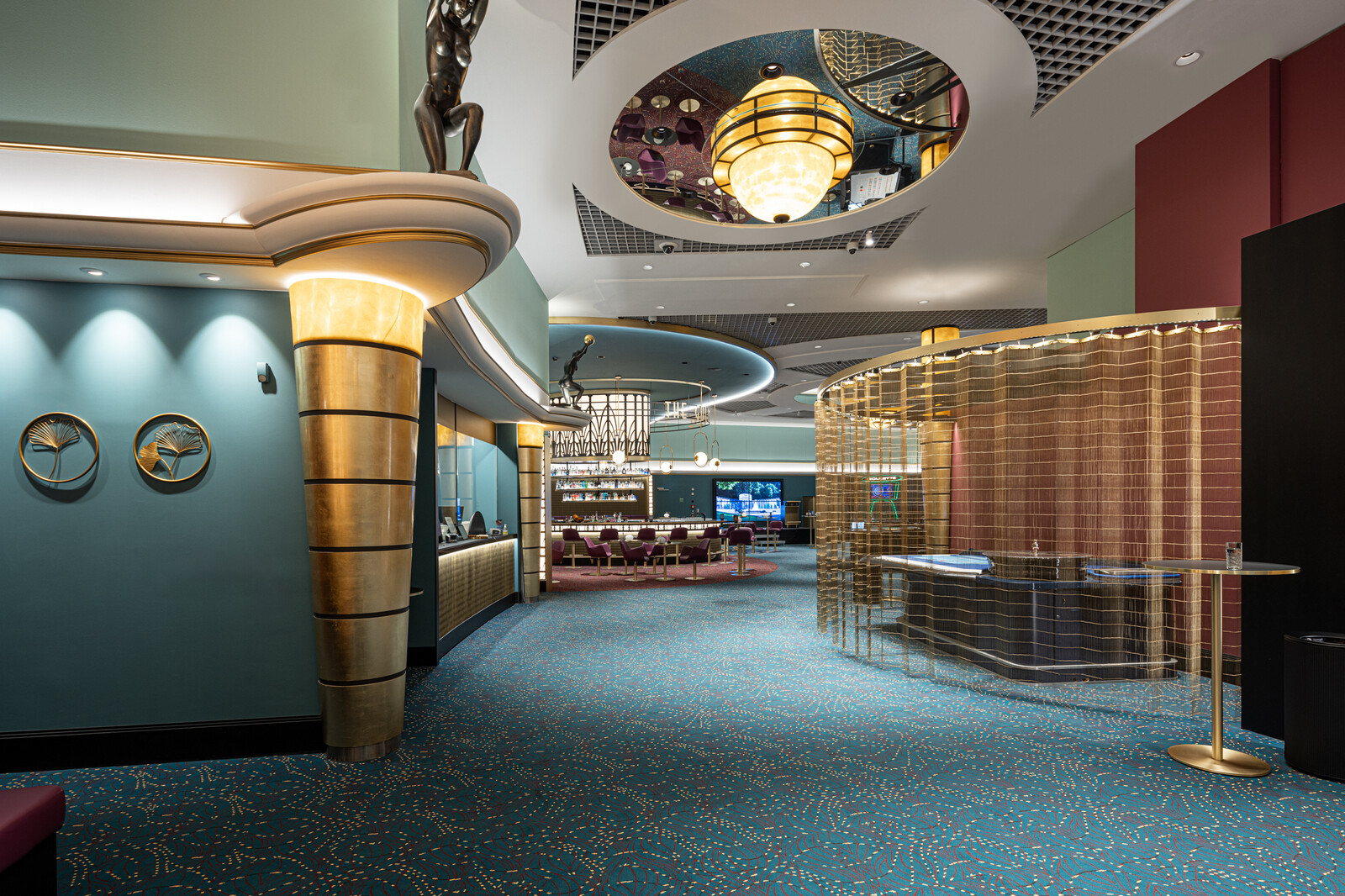 der Eingang zum Casino Bad Ragaz, mit tealfarbenem Teppich, goldenen Akzenten und moderner Beleuchtung. Ein geschwungener Tresen und plüschige Sitzgelegenheiten.