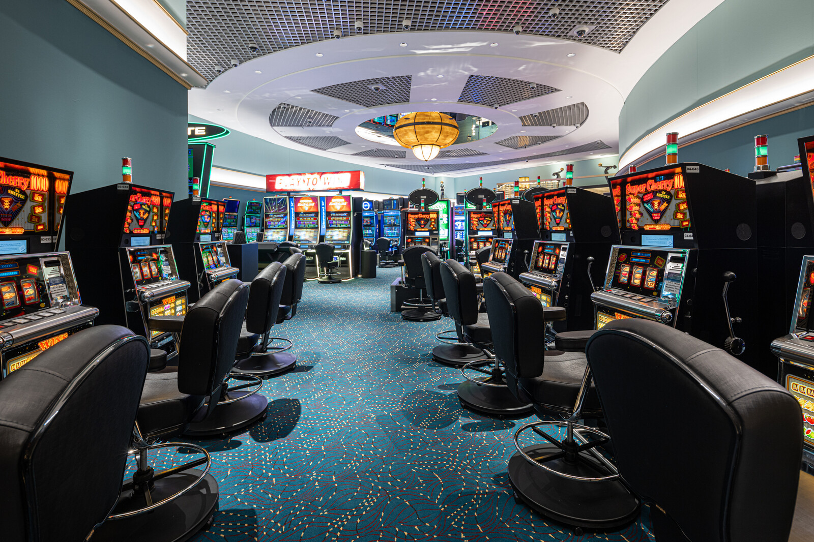 Ein hell erleuchteter Kasinosaal des Casino Bad Ragaz mit Reihen von bunten Spielautomaten und gepolsterten Hockern, mit einem üppigen Kronleuchter und einem gemusterten blauen Teppich.