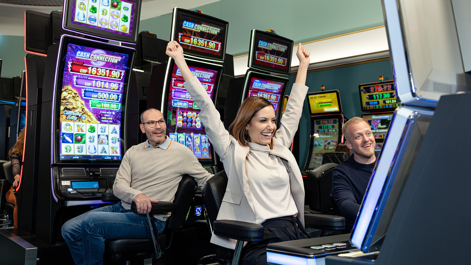 Eine Frau hebt aufgeregt die Arme, während sie an einem Spielautomaten sitzt, flankiert von zwei lächelnden Männern im Casino Bad Ragaz.