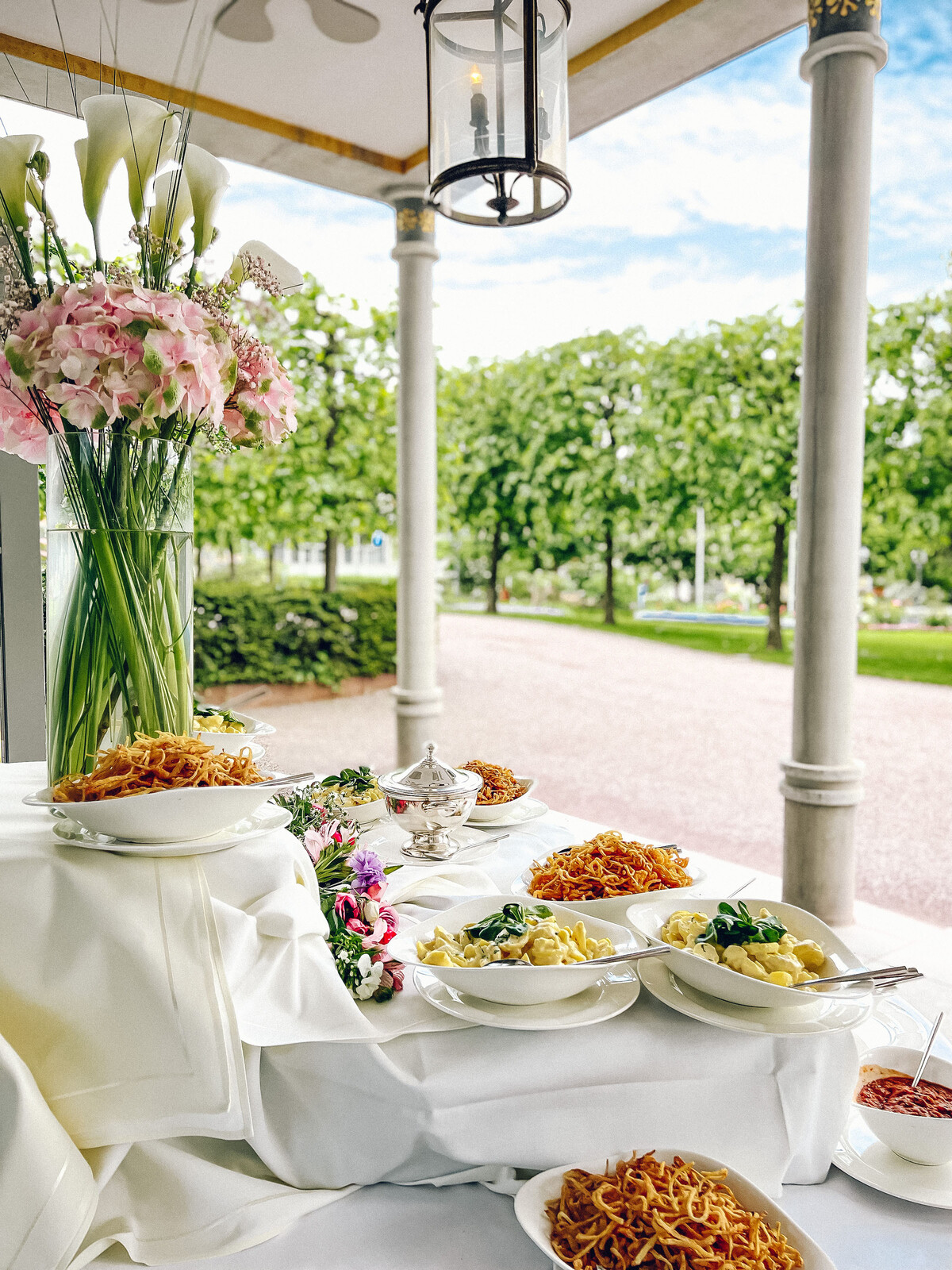Ein Tisch mit weissem Tischtuch steht auf der Terrasse des Restaurant Olives d'Or. Bäume reihen sich am Rand der Wiese des Parks entlang und die Sonne scheint auf das Buffet. Auf dem Tisch stehen verschiedene Speisen, unter anderem Gnocci und Zwiebeln. Eine Blumenvasen mit frischen rosanen und weissen Blumen zieren den Tisch. Das Restaurant Olives d'Or befindet sich in dem Wellnesshotel Grand Resort Bad Ragaz, einem Superior Luxushotel in der Schweiz.