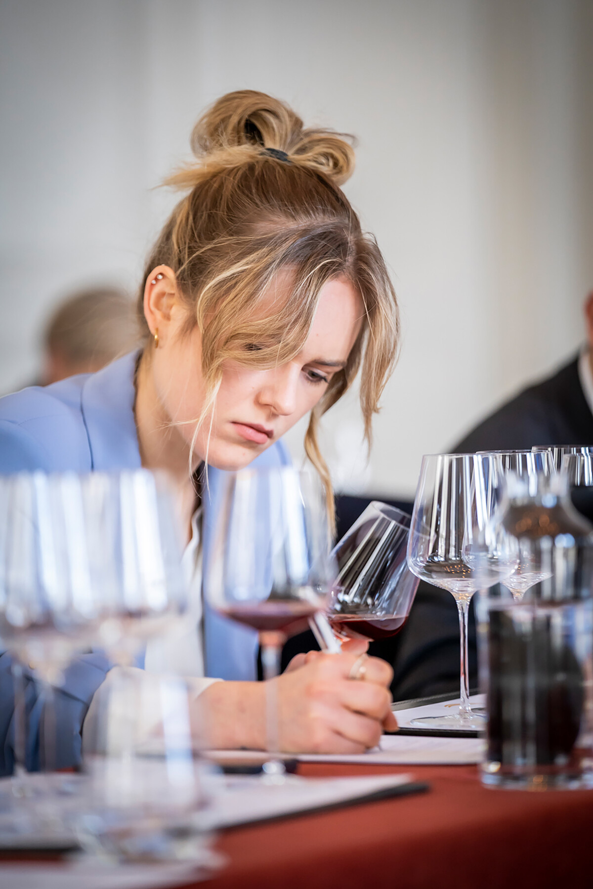 Eine junge Weinkennerin hält ein Glas gefüllt mit Rotwein in ihrer linken Hand. Mit der rechten Hand notiert sie etwas. Auf dem Tisch um sie herum stehen viele Weingläser.