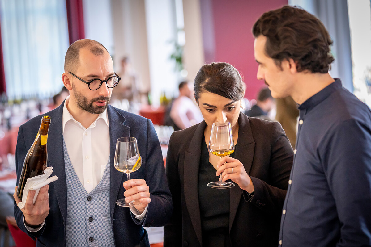 Drei Weinkenner stehen nebeneinander. In der Mitte steht eine Dame, die an einem Glas Weisswein riecht. Der Mann zu ihrer Rechten hält in seiner linken Hand ein Glas Weisswein und in seiner rechten Hand die dazugehörige Flasche mit einem Tuch zwischen seiner Hand und der Flasche. Der dritte Weinkenner steht links von der Frau. Er ist den beiden anderen interessiert zugewandt.
