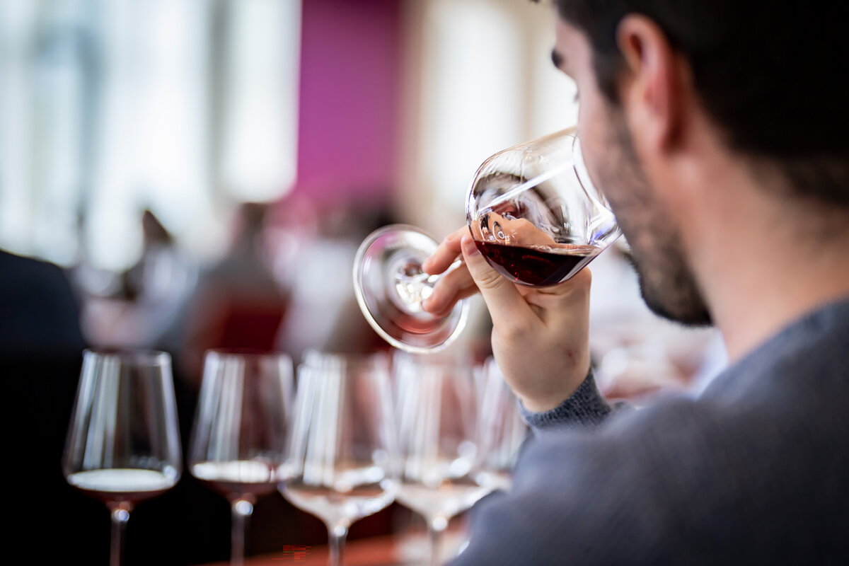 Blick über die Schulter eines jungen dunkelhaarigen Mannes. Er riecht an einem Glas Rotwein. Vor ihm sind weitere sieben Gläser Wein zu erahnen.
