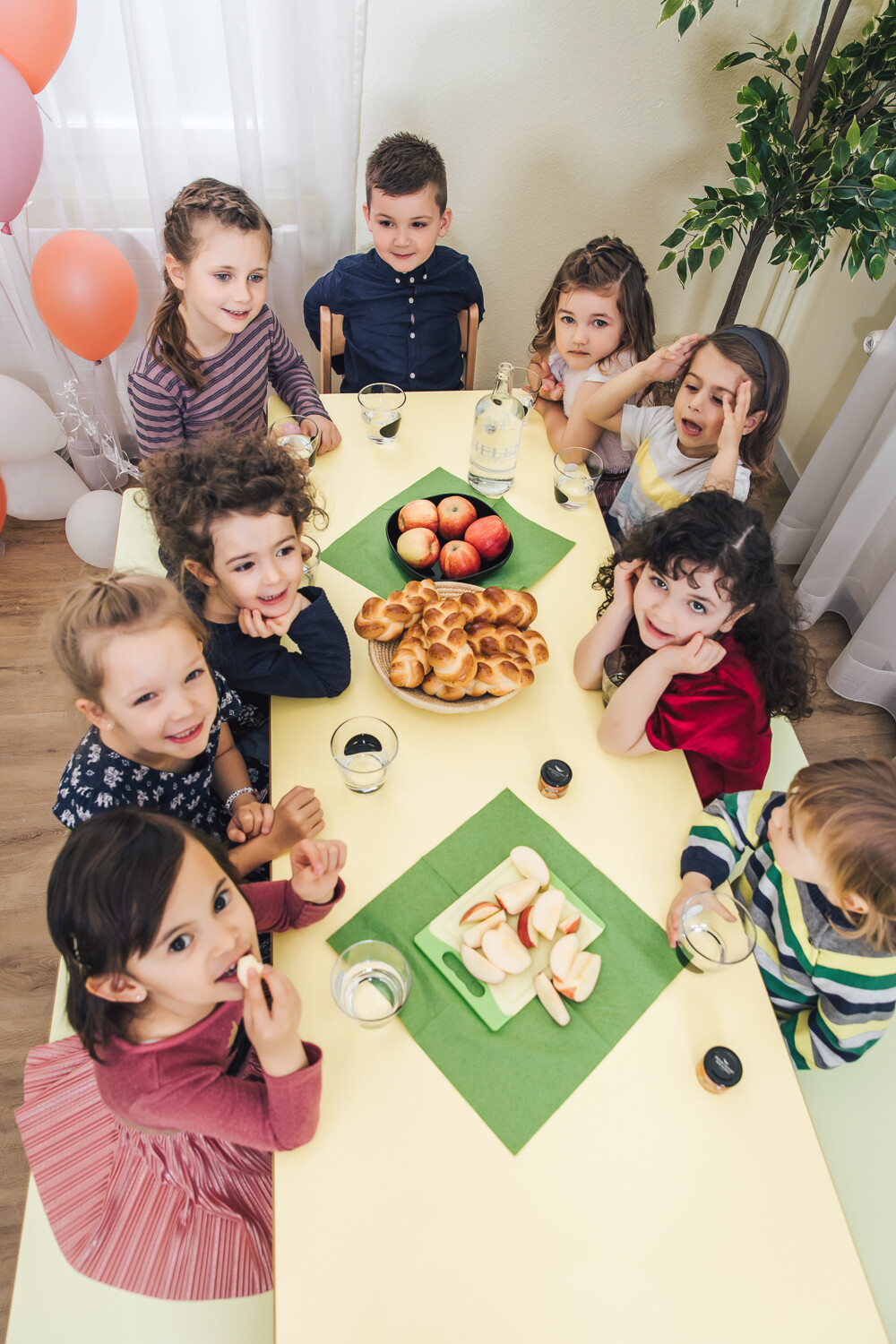 Eine Gruppe Kinder isst in der betreuten Kindervilla einen gesunden Snack. Dieser besteht aus Äpfeln und Brötchen. Sie lachen in die Kamera. Ein Mädchen in einem pinken Rock schiebt sich währenddessen einen Apfelschnitz in den Mund. In der oberen rechten Ecke befindet sich eine Pflanze und in der linken oberen Ecke schweben rosarote Luftballons. Die Kindervilla ist ein Teil des Wellnesshotel Grand Resort Bad Ragaz, einem Superior Luxushotel in der Schweiz.