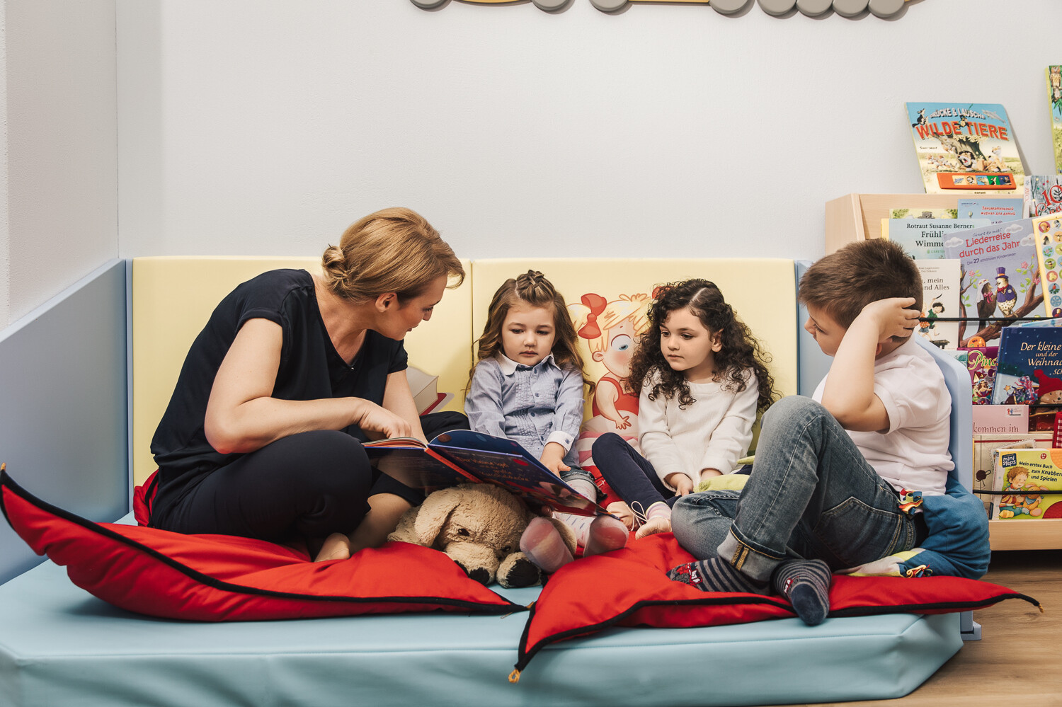 Eine Betreurin der Kindervilla sitzt mit drei Kinder, einem Jungen und zwei Mädchen, in einer gemütlichen Leseecke. Ein grosses Buch liegt auf dem Schoss des einen Mädchens und die Betreuerin liest ihr daraus vor. Die Kindervilla gehört zum Wellnesshotel Grand Resort Bad Ragaz, einem Superior Luxushotel in der Schweiz.