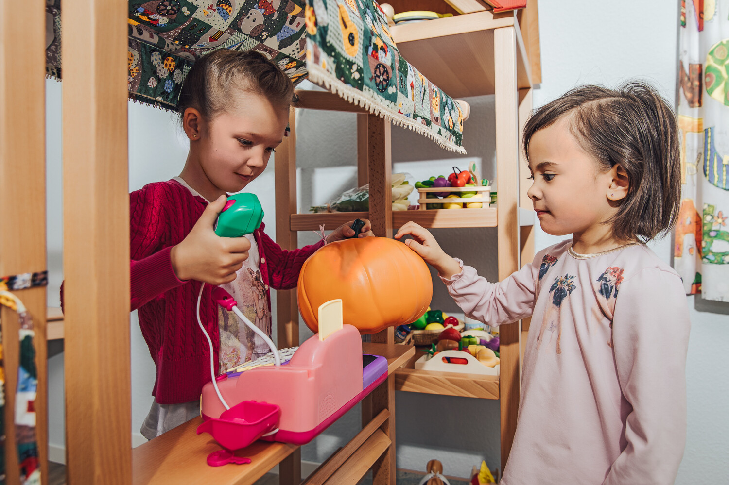 Zwei Kinder spielen in der Kindervilla mit einem Foodshop. Der Junge ist der Kassierer und scannt gerade einen Kürbis in die Kasse ein. Das Mädchen ist die Kundin und reicht ihm ihren Einkauf. Hinter den beiden befindet sich ein Holzregal, in dem sich noch mehr hölzerne Lebensmittel befinden. Die Kindervilla gehört zum Wellnesshotel Grand Resort Bad Ragaz, einem Superior Luxushotel in der Schweiz.