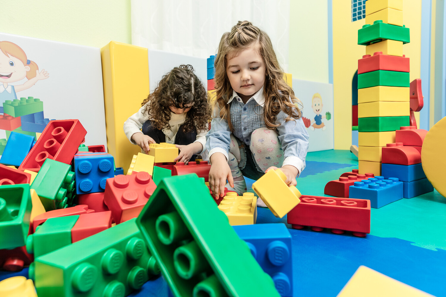 Riesige Legobausteine liegen auf dem Boden der Kindervilla. Zwei Mädchen spielen damit. Hinter ihnen türmt sich bereit ein riesiger Turm aus bunten Legobausteinen. Die Kindervilla gehört zum Wellnesshotel Grand Resort Bad Ragaz, einem Superior Luxushotel in der Schweiz.